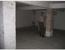 Eine leere Kellerhalle mit Betonboden und -decke, weißen und teils unverputzten Ziegelwänden. Eine stehende Person hält ein Werkzeug und lehnt an einer gemauerten Säule.