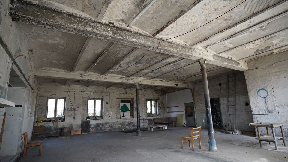 Großer, leerer Industriehallenraum mit freiliegenden, verwitterten Deckenbalken, abblätterndem Putz und mehreren Stahlstützen. Entlang der Backsteinwände befinden sich mehrere Fenster sowie vereinzelt abgestellte Möbel und eine Leiter auf dem Betonboden.