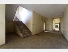 Ein leerer, heruntergekommener Innenraum mit sichtbaren Abnutzungsspuren. Links eine Treppe mit Metallgeländer, die nach oben führt, rechts ein langer Flur mit mehreren Türen und sichtbaren Durchblicken.