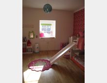 Kinderzimmer mit hellem Holzfußboden, einer weißen Rutsche, einem bunten Teppich in Blumenform und einem Bett. An der Wand befindet sich ein kleines Fenster, unter dem ein weißer Schrank steht, sowie eine rote Tapete mit Mustern auf der rechten Seite.