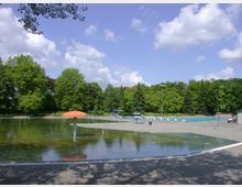 Ein Freibad mit einem flachen Wasserbereich im Vordergrund und einer orangefarbenen Pilz-Wasserspielstruktur. Im Hintergrund befinden sich ein tieferes Schwimmbecken, Liegeflächen, Bäume und eine grüne Parklandschaft unter blauem Himmel mit weißen Wolken.