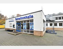 Außenansicht einer Schwimmhalle mit weißer Fassade, blauer Eingangstür und großen Fenstern mit Schwimmplakaten. Der Vorplatz ist mit Pflastersteinen ausgelegt, und rechts befinden sich blaue Fahrradständer.