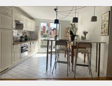 Helle Küche mit weißer Einbauküche, Holzarbeitsplatte und Fliesenboden. Im Vordergrund ein hoher Esstisch mit weißen Hockern, im Hintergrund eine Glastür, die zu einem Balkon führt, und dekorative Pflanzen sowie eine helle Wand mit einem Pflanzenposter.