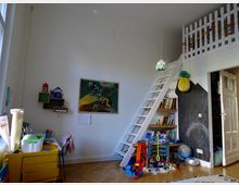 Kinderzimmer mit einem Hochbett, das über eine weiße Leiter erreichbar ist, und einem Holzgeländer. Links steht ein gelber Kinder-Schreibtisch, daneben eine Bildwand und schwebende Regale. Rechts gibt es ein Bücherregal, eine schwarze Tafelwand mit Zeichnungen und Spielsachen auf dem Boden. Holzboden und helle Wandgestaltung dominieren den Raum.