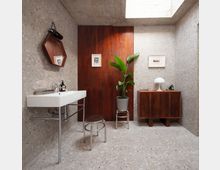 Modern bathroom with terrazzo walls and floor, a white wall-mounted sink, and a central wood-paneled partition. A skylight brightens the space, which includes a wooden cabinet with a table lamp, framed artwork, a round stool, and a potted plant.