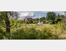 Ländliche Szene mit Wiesen und Büschen im Vordergrund, im Hintergrund eine kleine Ortschaft mit Häusern und einem Gebäude mit rotem Dach und einem Turm, umgeben von Bäumen. Der Himmel ist leicht bewölkt.