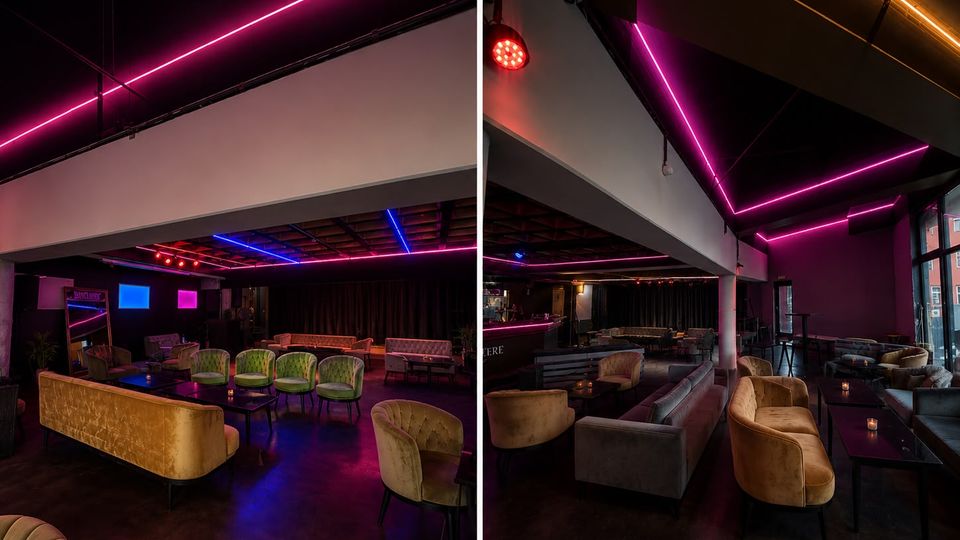 Innenraum einer modernen Lounge- oder Bar mit niedriger Decke und farbigen Neon-Lichtleisten in Pink, Blau und Orange. Samtsofas und gepolsterte Sessel gruppieren sich um niedrige Tische, im Hintergrund sind eine Bar und ein abgetrennter Bühnen- oder Vorhangbereich zu sehen.