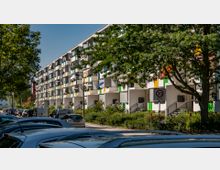 Mehrstöckiges Wohngebäude mit modernen, farbigen Balkonen und einer Grünanlage im Vordergrund. Geparkte Autos säumen die Straße, und ein Schild weist auf eine Tempo-30-Zone hin.