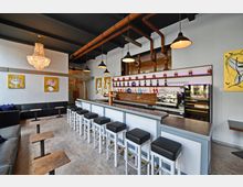 Barbereich eines modernen Cafés mit heller Einrichtung. Eine lange Theke mit Barhockern steht zentral im Raum, dahinter befinden sich Regale mit Flaschen, eine Kaffeemaschine und eine Vitrine. Die Wände sind mit gelben Kunstbildern verziert, und über der Sitzlounge hängt ein großer Kronleuchter.