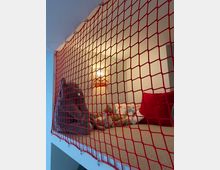 Eine erhöhte Spielecke mit Teppichboden, umgeben von einem roten Sicherheitsnetz. Im Hintergrund befinden sich Kuscheltiere, Kissen und eine Wandleuchte, die den Bereich beleuchtet.