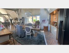 Ein stilvoll eingerichtetes Wohnzimmer mit offenem Dachstuhl, einem großen Esstisch aus Holz, gepolsterten Stühlen und einem Teppich. Im Raum befinden sich eine Glasvitrine, ein Kronleuchter, dekorative Möbel sowie ein Kamin im Hintergrund.