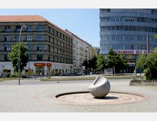 Städtischer Platz mit einer runden Stein-Skulptur in der Mitte, umgeben von gepflastertem Boden und niedrigen Bäumen. Im Hintergrund befinden sich ein mehrstöckiges, historisches Gebäude mit einem Penny-Markt im Erdgeschoss sowie ein modernes Bürogebäude.
