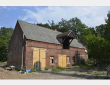 Backstein-Gebäude mit verwitterter Fassade und wellblechartigem Dach, umgeben von Bäumen. Einige Fenster und Türen sind mit Holzplatten verbarrikadiert, während der Eingangsbereich unbewirtschaftet wirkt.