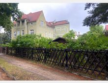 Grüne Wohngegend mit einem großen, historischen Mehrfamilienhaus in hellgrüner Farbe und roten Ziegeldächern, verziert mit Stuck-Elementen. Im Vordergrund ein Holzzaun und dichter Bewuchs, daneben ein kleiner Weg.