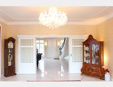 Innenraum eines eleganten Wohnbereichs mit Marmorboden, einer verzierten weißen Schiebetür mit Glasfenstern, einem Kronleuchter und gelbem Tapetenmuster. Links steht eine Standuhr aus Holz, rechts eine antike Vitrine mit Porzellan. Im Hintergrund sind eine weiße Tür, große Fenster und eine geschwungene Treppe zu sehen.