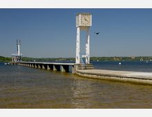 Eine Anlegestelle am Wasser mit einem langen Steg, der ins See führt. Im Vordergrund steht ein weißer Uhrturm mit kreisförmigen Aussparungen, und im Hintergrund sind Bäume sowie einige Gebäude am Ufer sichtbar.