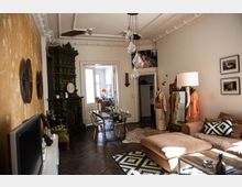 Wohnzimmer in einem Altbau mit hohen Decken, Stuckverzierungen und Holzboden. Der Raum enthält einen großen grünen Kachelofen, eine Sitzecke mit beigem Sofa, einen Esstisch mit Stühlen sowie dekorative Elemente wie einen großen vergoldeten Spiegel, Wandkunst und hängende Glühbirnenlampen.