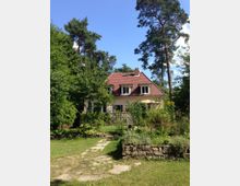 Einfamilienhaus mit rotem Ziegeldach und cremefarbener Fassade, umgeben von einem großzügigen Garten mit blühenden Pflanzen, gepflegtem Rasen und einem gepflasterten Weg. Im Hintergrund stehen hohe, schlanke Kiefern.