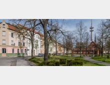 Straßenansicht mit Wohnhäusern in Pastelltönen, kleinen Vorgärten und parkenden Autos. Im Vordergrund ein begrüntes Parkgelände mit Bäumen, Gehwegen und Sitzbänken, daneben ein Backsteingebäude und ein hoher Funkmast.