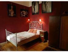 Schlafzimmer mit dunkelroten Wänden, einem Bett mit weißem Metallrahmen und heller Bettwäsche sowie zwei Nachttischlampen an der Wand. An den Wänden hängen mehrere Fotos, und rechts steht ein Holzkleiderschrank.