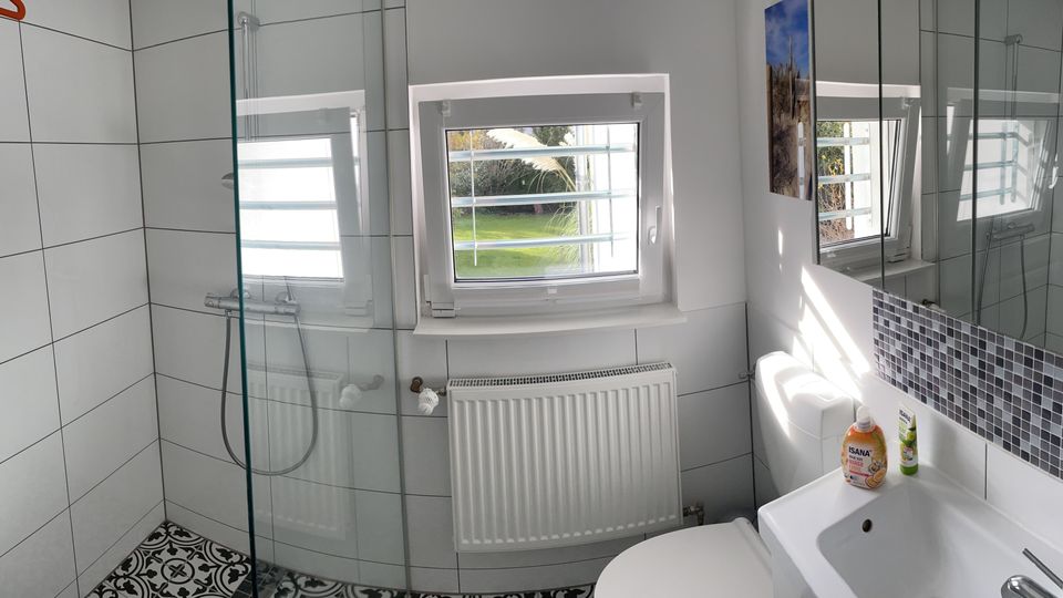 Helles, gefliestes Badezimmer mit begehbarer Glasdusche, kleinem Fenster über einem Heizkörper und einem Waschtisch mit großem Spiegel und Mosaikfliesenbordüre. Schwarz-weiß gemusterte Bodenfliesen ergänzen die weißen Wandfliesen.
