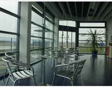 Innenraum eines modernen Cafés mit großen Glasfenstern, die den Blick auf einen Außenbereich mit Zaun und freiem Feld ermöglichen. Der Raum ist mit silbernen Metallstühlen, runden Glastischen und einer großen Zimmerpflanze eingerichtet.