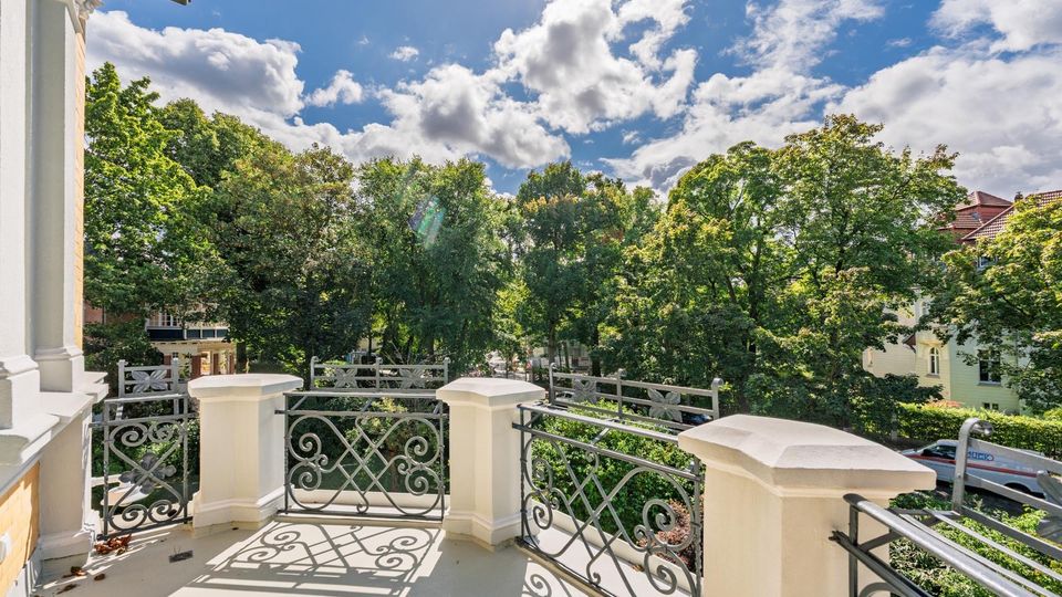 Balkon eines Wohnhauses mit hellen Pfeilern und dekorativem schmiedeeisernem Geländer, mit Blick auf eine von hohen Bäumen gesäumte Wohnstraße. Im Hintergrund sind weitere Häuserdächer unter blauem Himmel mit Wolken zu sehen.