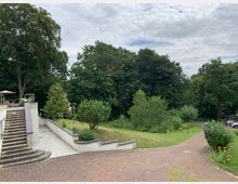Weitläufiger Garten mit einer gepflasterten Terrasse und angelegten Grünflächen, umgeben von großen Bäumen. Rechts führt ein geschwungener Weg durch die Wiese, während links eine Treppe zu einem erhöhten Bereich mit Sitzmöglichkeiten führt.