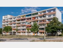 Ein mehrstöckiges Wohngebäude mit bunten Balkonen und Markisen, umgeben von einigen Bäumen und einer Rasenfläche. Der Himmel ist klar mit wenigen Wolken, eine Laterne steht im Vordergrund.