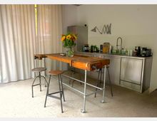 Helle Küche mit einer Metallarbeitsfläche, einem Waschbecken und diversen Küchenutensilien an der Rückwand. Im Vordergrund steht ein Tisch aus Holz und Metallrohren mit Barhockern darunter, dekoriert mit einem Blumenstrauß in einer Glasvase.