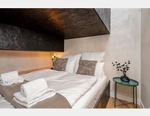 Modernes Schlafzimmer mit Doppelbett und weißer Bettwäsche unter einer markanten, dunklen, kantigen Deckenverkleidung. Neben dem Bett steht ein kleiner grüner Beistelltisch mit Vase, dazu eine schlichte Wandlampe und helle, glatte Wände.