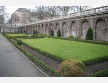 Ein gepflegter Garten mit rechteckigen Rasenflächen, umgeben von niedrigen Hecken und geformten Sträuchern, grenzt an eine lange, helle Steinmauer mit gewölbten Nischen, die von Kletterpflanzen bewachsen sind. Im Hintergrund sind mehrstöckige Wohngebäude und kahle Bäume zu sehen.