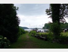 Grünfläche mit Bänken führt zu einem kleinen Hafenbereich; im Hintergrund sind Boote, Segelmasten und ein Wald am Seeufer zu sehen. Der Himmel ist bewölkt, die Uferzone ist von dichtem Grün und Bäumen eingerahmt.