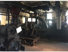 Innenraum einer alten Fabrikhalle mit historischen Maschinen und großen, dunklen Metallteilen wie Zahnrädern, Kesseln und Rohrleitungen. Die Umgebung wirkt industriell mit sichtbaren Stahlträgern, Fenstern im Hintergrund und einer gedämpften Beleuchtung.