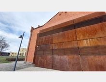 Backsteinfassade eines Gebäudes mit großen, rostfarbenen Metallplatten an einer Seitenwand. Im Vordergrund ein gepflasterter Gehweg und eine Straßenlaterne, im Hintergrund sind weitere Gebäude und eine Grünfläche zu sehen.