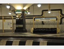 Bahnhof der Berliner U-Bahn-Linie U7 mit gelb-schwarzen Dekorationselementen und einer Beschilderung für "Rathaus Spandau". Sitzbänke, eine zentrale Säule und helle Deckenleuchten prägen die Szenerie.