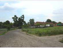 Ein ländlicher Weg aus festgetretener Erde führt an Weiden mit grünen Zäunen entlang. Im Hintergrund stehen Bäume und ein rotes Backsteingebäude mit landwirtschaftlichen Geräten und Anhängern auf einer Wiese.