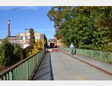 Eine schmale Fußgängerbrücke mit grünen Geländern führt zu einem Industriegelände mit Backstein- und hellen Fassaden. Im Hintergrund sind ein rauchloser Schornstein, ein roter Lastwagen und dichte Bäume zu sehen; ein Mann mit Hund geht auf der Brücke entlang.