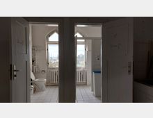 Zwei offene Toilettentüren in einem schlicht gestalteten Badezimmer mit weißen Fliesen, sichtbaren Rohren und einem Heizkörper unter gotisch geformten Fenstern. Durch die Fenster ist eine städtische Aussicht mit Gebäuden und Schornsteinen zu sehen.