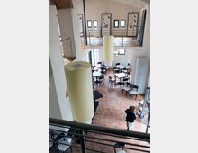 Innenraum eines zweistöckigen Gebäudes mit einem offenen, hellen Bereich. Im unteren Stockwerk befinden sich Terrakottaboden, mehrere Tische und Stühle sowie große, zylindrische Hängelampen, während das obere Geschoss eine Galerie mit Geländern und Plakaten an den Wänden zeigt.