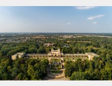 Luftaufnahme eines historischen Palastkomplexes mit symmetrischer Gartenanlage, umgeben von dichtem Wald. Im Hintergrund sind ein See, Wohnhäuser und weite Landschaft zu sehen.