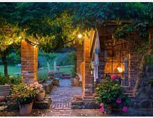 Beleuchteter Gartenbereich mit Ziegelpfeilern und einer Pergola, die von Rankpflanzen bedeckt ist. Im Hintergrund stehen Blumen und Pflanzen in Blumentöpfen, während ein großer Fensterbereich eines Gebäudes mit warmem Innenlicht sichtbar ist.
