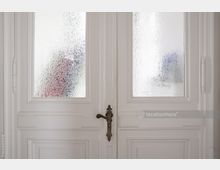 Weiße zweiflügelige Tür mit satinierten Glaseinsätzen, verziertem Messinggriff und klassischem Rahmenprofil. Im Hintergrund sind undeutlich farbige Formen durch das strukturierte Glas erkennbar.