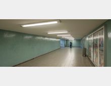 Unterführung mit hellgrünen Kachelwänden, einer gefliesten Bodenfläche und einer Decke mit mehreren Leuchtstofflampen. Rechts befinden sich Werbeposter hinter Glas, links eine leere Wand, und im Hintergrund steht eine Person nahe einem Ausgang mit Treppen.