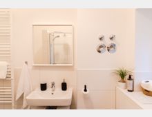 Helles, modernes Badezimmer mit Waschbecken, Spiegel, weißem Handtuchhalter und Accessoires wie Seifenspender und Zahnputzbecher. Rechts befinden sich eine kleine Pflanze, ein Toilettenpapierhalter sowie ein Korb mit Toilettenpapierrollen.