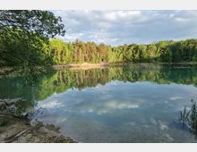 Ein klarer See mit ruhiger Wasseroberfläche, die den Himmel und umliegende Bäume spiegelt. Im Hintergrund dichter Wald, während im Vordergrund ein sandiges Ufer mit Gräsern zu sehen ist.