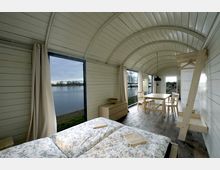 Innenraum einer gemütlichen Holzhütte mit gewölbter Decke und großer Fensterfront, die den Blick auf einen Fluss und eine Landschaft mit Bäumen ermöglicht. Der Raum ist mit einem Doppelbett, einer Kommode, einem Esstisch mit Stühlen und Holzmöbeln minimalistisch eingerichtet.