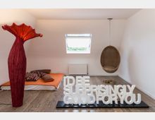 Minimalistisch eingerichteter Raum mit einem niedrigen Bett mit orangefarbener Decke und bunten Kissen, einer hängenden Sitzschale aus Rattan, einer großen roten Lampe mit skulpturalem Design und einem weißen Schriftzug aus 3D-Buchstaben auf dem Boden. Im Hintergrund befindet sich ein schräges Dachfenster mit Blick nach draußen.