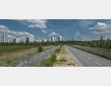 Straßenansicht mit einer asphaltierten Fahrbahn rechts und einer umzäunten Grünfläche links, im Hintergrund mehrere mehrstöckige Wohngebäude und Baumgruppen unter einem wolkigen blauen Himmel.