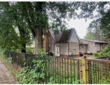 Ein älteres Gebäude mit verwittertem Putz und einem roten Ziegeldach, teilweise überwuchert von Grünpflanzen. Vor dem Haus steht ein hoher Baum, daneben ein Metallzaun, und im Hintergrund sind weitere Vegetation und Nebengebäude erkennbar.